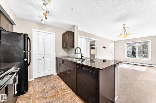 Kitchen island with dark cabinetry, integrated dishwasher, and undermount sink - #218 5810 Mullen Pl Nw Nw, Edmonton, AB 