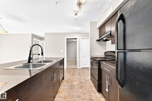 Kitchen featuring a double basin stainless steel sink, black gooseneck faucet, dark cabinetry, and tile flooring - #218 5810 Mullen Pl Nw Nw, Edmonton, AB 