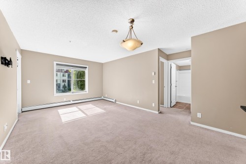 Neutral wall color, carpeted flooring, window with white trim, built-in baseboard heating, and an overhead decorative light fixture - #218 5810 Mullen Pl Nw Nw, Edmonton, AB 