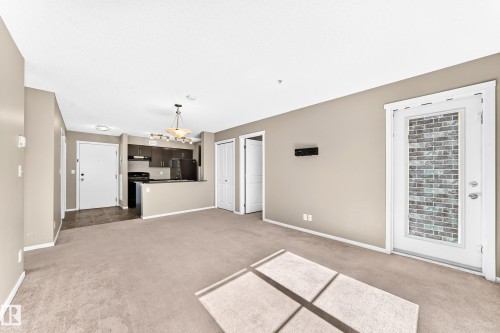 Open-concept living space with carpeting, kitchen with dark wood-finish cabinetry, black appliances, and a tiled entry - #218 5810 Mullen Pl Nw Nw, Edmonton, AB 