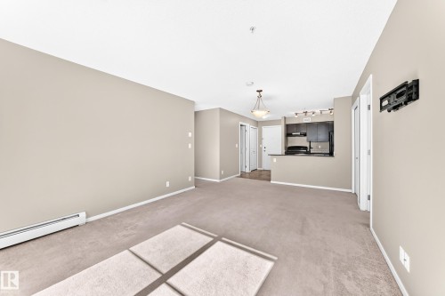 Open-concept living area featuring neutral carpeting and walls, overhead lighting, and built-in kitchen cabinetry - #218 5810 Mullen Pl Nw Nw, Edmonton, AB 