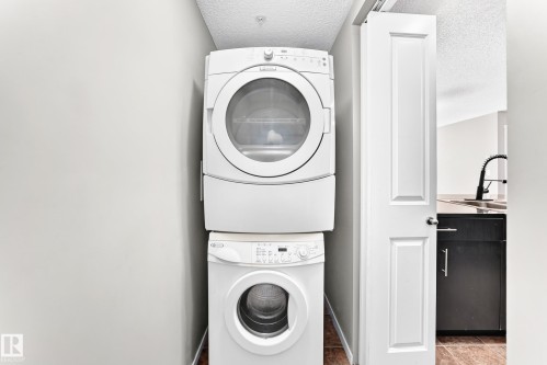 Stacked washer and dryer unit with a paneled door, black cabinetry, and a gooseneck faucet - #218 5810 Mullen Pl Nw Nw, Edmonton, AB 