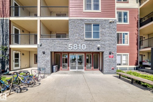 Building exterior featuring stone cladding, multi-story balconies with black railings, a ground-level entrance with glass doors, and an integrated bicycle rack - #218 5810 Mullen Pl Nw Nw, Edmonton, AB 