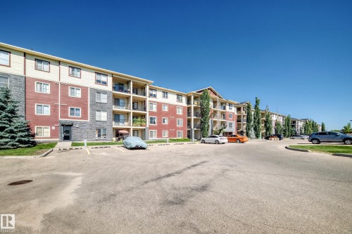 Multi-story building featuring balconies, red and grey siding, and ample surface parking - #218 5810 Mullen Pl Nw Nw, Edmonton, AB 