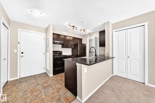 Kitchen featuring dark wood-finish cabinetry, black appliances, a black countertop, a pull-down faucet, and an island - #218 5810 Mullen Pl Nw Nw, Edmonton, AB 
