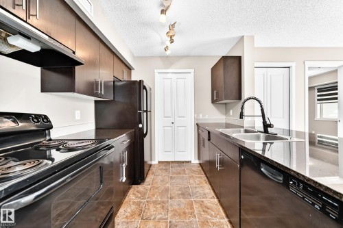 Kitchen featuring wood-finish cabinetry, dark countertops, tile flooring, a double basin sink with a gooseneck faucet, and stainless steel appliances - #218 5810 Mullen Pl Nw Nw, Edmonton, AB 