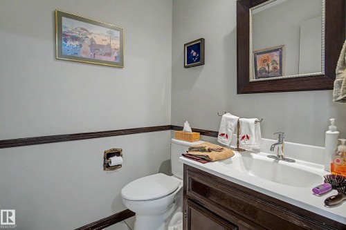 White-painted walls with dark wood trim, a white ceramic toilet, a dark wood-finish vanity with a white countertop, a polished chrome faucet, and a framed mirror - 36 Lodgepole Crescent, St. Albert, AB 