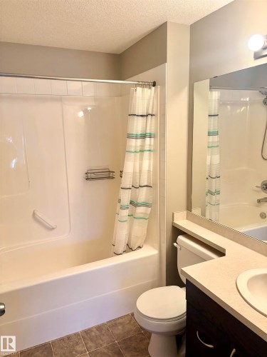 White bathtub with shower, dark wood-finish vanity, light-toned countertop, integrated sink, and large wall-mounted mirror - #1304 9363 Simpson Dr Nw, Edmonton, AB 