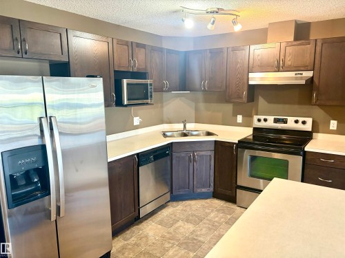Kitchen with wood-finish cabinetry, stainless steel appliances, light countertops, a double basin sink, and track lighting - #1304 9363 Simpson Dr Nw, Edmonton, AB 