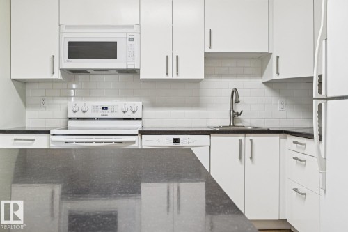 White cabinetry, dark stone countertops, white subway tile backsplash, stainless steel sink with gooseneck faucet, and white appliances - #905 9929 113 St Nw, Edmonton, AB 