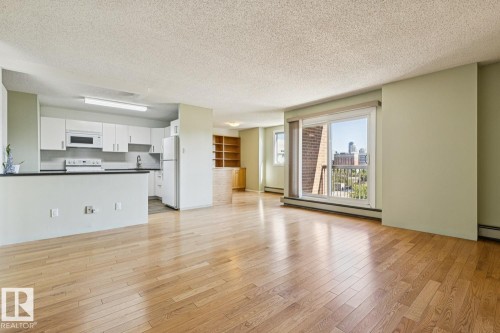 Open-concept living space featuring wood-finish flooring, a kitchen with white cabinetry and dark countertops, built-in shelving, and a sliding glass door leading to a balcony - #905 9929 113 St Nw, Edmonton, AB 