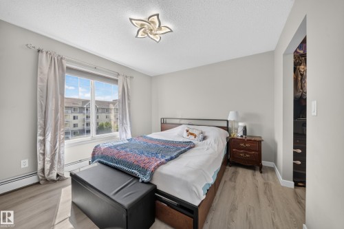 Bright, naturally lit bedroom featuring hardwood flooring, a large window with thermal curtains, a wood-finish nightstand, and a decorative ceiling fixture - #419 4407 23 St Nw, Edmonton, AB 
