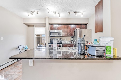 Kitchen featuring dark wood cabinetry, stainless steel appliances, a double basin sink, track lighting, and a dark stone-finish countertop - #225 111 Watt Cm Sw, Edmonton, AB 