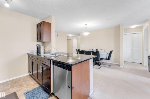 Kitchen island featuring a double basin sink, stainless steel dishwasher, dark cabinetry, and a dark countertop - #225 111 Watt Cm Sw, Edmonton, AB 