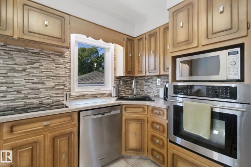 Wood-finish cabinetry paired with light-toned countertops, a mosaic tile backsplash, stainless steel appliances, and a built-in oven and microwave - 12916 63 Street Nw, Edmonton, AB 