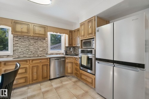 Kitchen featuring wood cabinetry, stainless steel appliances, tile backsplash, light-toned countertops, and ceramic tile flooring - 12916 63 Street Nw, Edmonton, AB 