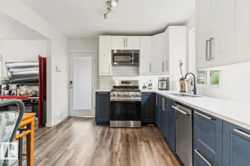 Kitchen featuring two-tone cabinetry with white upper and blue lower cabinets, stainless steel appliances, wood-finish flooring, white countertops, and a white subway tile backsplash - 12150 47 Street Nw, Edmonton, AB - Indoor Photo Showing Kitchen