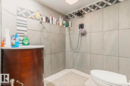 Bathroom featuring a curved wood-finish vanity with a white countertop, light gray square wall tiling, and a mosaic tile border - 12150 47 Street Nw, Edmonton, AB - Indoor Photo Showing Bathroom