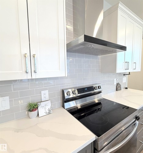 White cabinetry with metal hardware, light-toned subway tile backsplash, stainless steel range with a black cooktop, stainless steel range hood, and light-toned countertops - 1003 Goldfinch Way Nw, Edmonton, AB 