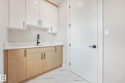 Modern kitchenette featuring warm wood-finish cabinetry with integrated hardware, white upper cabinetry with glass fronts, marble countertops, and a matte black single-handle faucet - 16 Middleton Close, Leduc, AB 