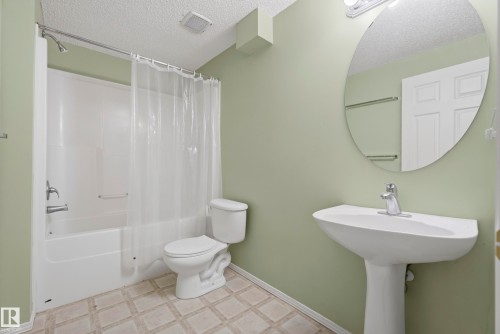 Pedestal sink with chrome faucet, circular wall mirror, and a white bathtub with shower fixtures, set on square-patterned flooring - 15213 49A Street Nw, Edmonton, AB 