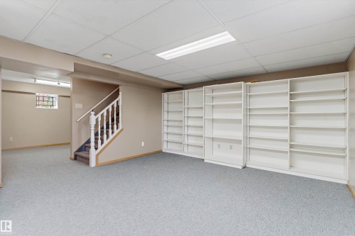 Expansive room featuring wall-to-wall carpeting, extensive built-in shelving, a drop ceiling with integrated lighting, and a staircase with white balusters - 5501 48 Avenue, Wetaskiwin, AB 