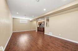 Expansive open-concept room featuring warm wood-finish flooring, recessed lighting, a neutral wall palette, and a partial wall opening to a kitchen area with dark wood cabinetry and stone-look countertops - 
