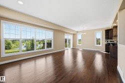 Spacious living area featuring expansive windows with scenic water and greenery, rich wood-finish flooring, a sliding glass door to an exterior balcony, recessed ceiling lighting, and an adjacent kitchen area with dark cabinetry - 