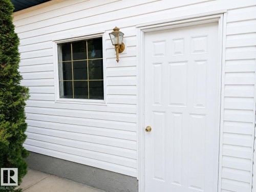White paneled exterior with a single window featuring grid muntins, an exterior wall sconce, and a white paneled door with a brass-finish doorknob - 3525 22 St Nw, Edmonton, AB 