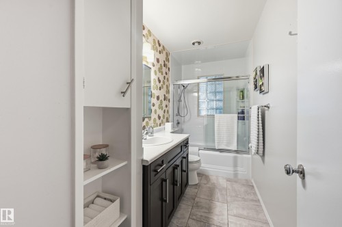 Full bathroom featuring a dark wood-finish vanity with an integrated sink, a glass-enclosed shower-over-tub with glass block accents, and tile flooring - 10552 45 Street Nw, Edmonton, AB 