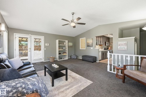Vaulted ceiling living area with a ceiling fan light fixture, double French doors, and built-in shelving, opening to a kitchen with gray cabinetry and stainless steel appliances - 10552 45 Street Nw, Edmonton, AB 