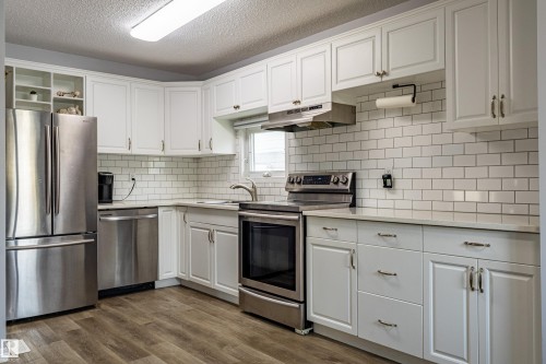 White shaker-style cabinetry with gold-tone hardware, stainless steel appliances, subway tile backsplash, light-toned countertops, and wood-finish flooring - 7408 180 Street Nw, Edmonton, AB 
