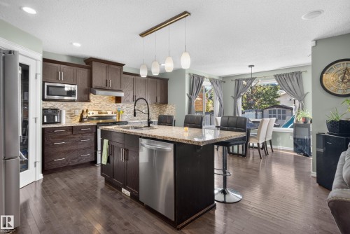 Kitchen featuring dark wood cabinetry, stainless steel appliances, a stone countertop island with seating, a tile backsplash, and recessed lighting - 1332 72 Street Sw, Edmonton, AB 