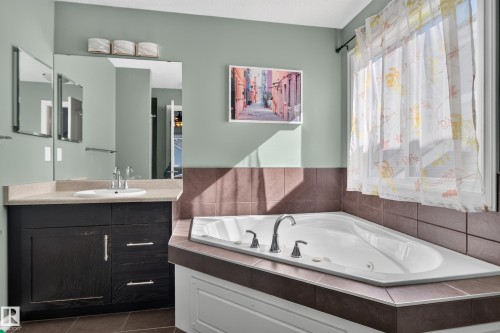 Spa-inspired primary bathroom showcasing a sunken soaking tub with dual faucet fixtures, custom tile surround in warm brown tones, and an expansive vanity with dark cabinetry and quartz countertop - 1332 72 Street Sw, Edmonton, AB 
