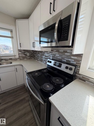 Modern kitchen featuring white cabinetry with black hardware, light-toned countertops, a glass tile backsplash, stainless steel microwave, and wood-finish flooring - 7604 7606 75 Street Nw, Edmonton, AB - Indoor Photo Showing Kitchen