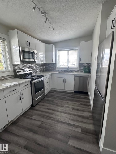 U-shaped kitchen featuring white cabinetry, stainless steel appliances, a tile backsplash, light countertops, and wood-finish flooring - 7604 7606 75 Street Nw, Edmonton, AB - Indoor Photo Showing Kitchen With Upgraded Kitchen