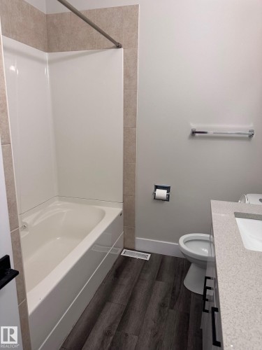 Bathroom featuring a white tub/shower combination with beige tile surround, light gray walls, dark wood-finish flooring, a white toilet, and a light-colored countertop with an integrated sink - 7604 7606 75 Street Nw, Edmonton, AB - Indoor Photo Showing Bathroom