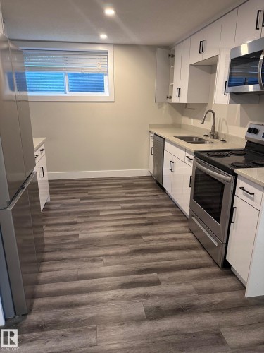 Kitchen featuring wood-finish flooring, white cabinetry with black hardware, stainless steel appliances, light countertops, and recessed lighting - 7604 7606 75 Street Nw, Edmonton, AB - Indoor Photo Showing Kitchen With Double Sink