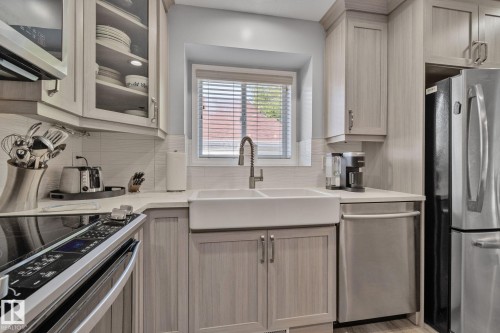 Light-toned cabinetry with glass-front accents, white countertop, farmhouse sink with a pull-down faucet, stainless steel appliances, and a tiled backsplash - 18812 89 Avenue Nw, Edmonton, AB 
