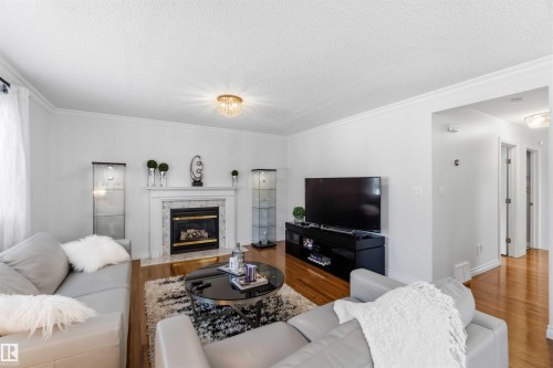 Living area featuring wood-finish flooring, a fireplace with a stone surround and mantel, crown molding, and a decorative ceiling fixture - 522 Klarvatten Lake Wynd Nw, Edmonton, AB 