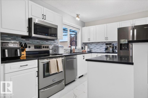 White cabinetry with contrasting black hardware, stainless steel appliances, dark countertops, a tiled backsplash, and a central island - 1217 76 Street Sw, Edmonton, AB 