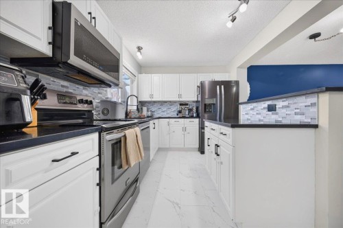 White cabinetry with matte black hardware, stainless steel appliances, black countertops, a mosaic tile backsplash, and white tile flooring - 1217 76 Street Sw, Edmonton, AB 