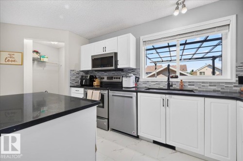 Kitchen featuring white cabinetry, dark countertops, stainless steel appliances, a tiled backsplash, and a window above the sink - 1217 76 Street Sw, Edmonton, AB 