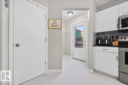 White paneled cabinetry with dark hardware, dark countertops, a tiled backsplash, stainless steel appliances, and white tile flooring - 1217 76 Street Sw, Edmonton, AB 