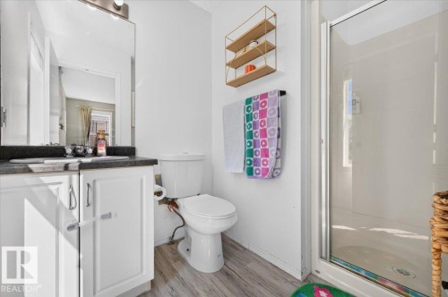 Wood-finish flooring, white vanity with dark countertop, large mirror with vanity lighting, standing shower with glass enclosure, wall-mounted shelving - 1217 76 Street Sw, Edmonton, AB 
