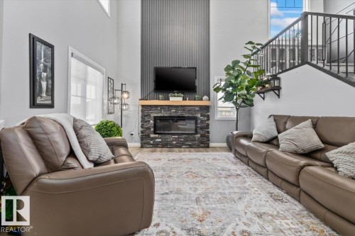 Living area featuring a floor-to-ceiling stone-clad fireplace with wood mantel, a distinctive fluted accent wall, wood-finish flooring, and a contemporary open-riser staircase with black metal railings - 5203 23A Avenue Sw, Edmonton, AB - Indoor Photo Showing Living Room With Fireplace