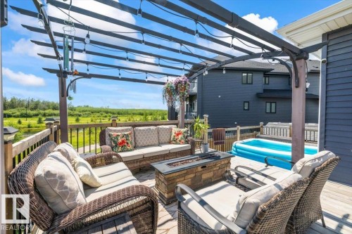 Expansive outdoor deck featuring a dark wood pergola with string lighting, a stone-finish fire pit, and a dark wood railing overlooking a natural landscape - 5203 23A Avenue Sw, Edmonton, AB - Outdoor With Deck Patio Veranda