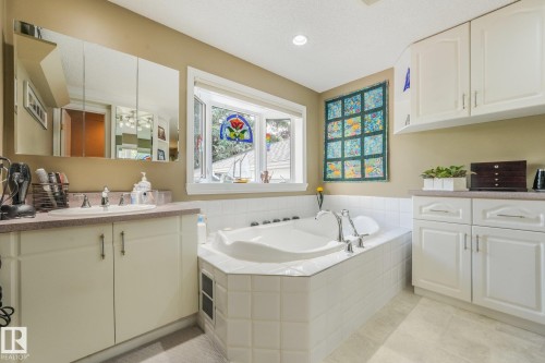 Built-in corner soaking tub, white vanity with integrated sink, white storage cabinetry, tiled flooring, and a bay window overlooking the rear garden. - 10531 137 Street Nw, Edmonton, AB 