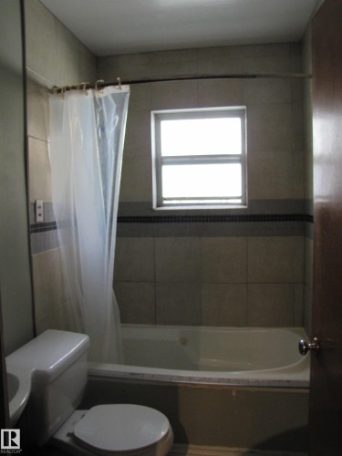 Tiled shower/tub combination with a window, dark accent tile band, and a white toilet - 9512 63 Avenue Nw, Edmonton, AB 