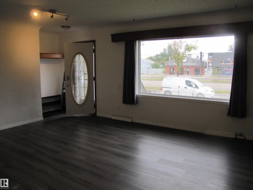 Spacious living area featuring wood-finish flooring, a large picture window with dark drapes, and a decorative front door with an oval glass insert - 9512 63 Avenue Nw, Edmonton, AB 
